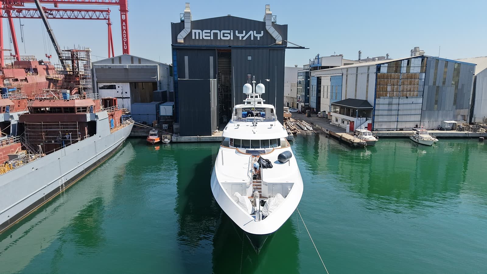 A motor yacht lying bow-on to the covered hall at the Mengi Yay yard in Tuzla, with a part-built commercial hull and a shipyard gantry crane on the quay alongside
