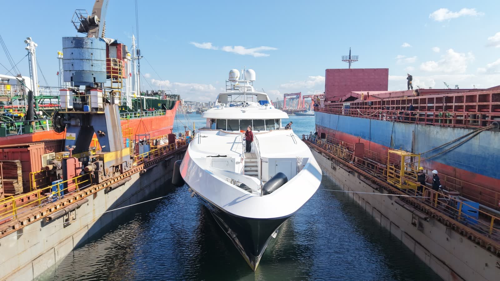 A superyacht in a floating dock in the Tuzla shipyard district, flanked by two commercial hulls under repair, with yard cranes behind