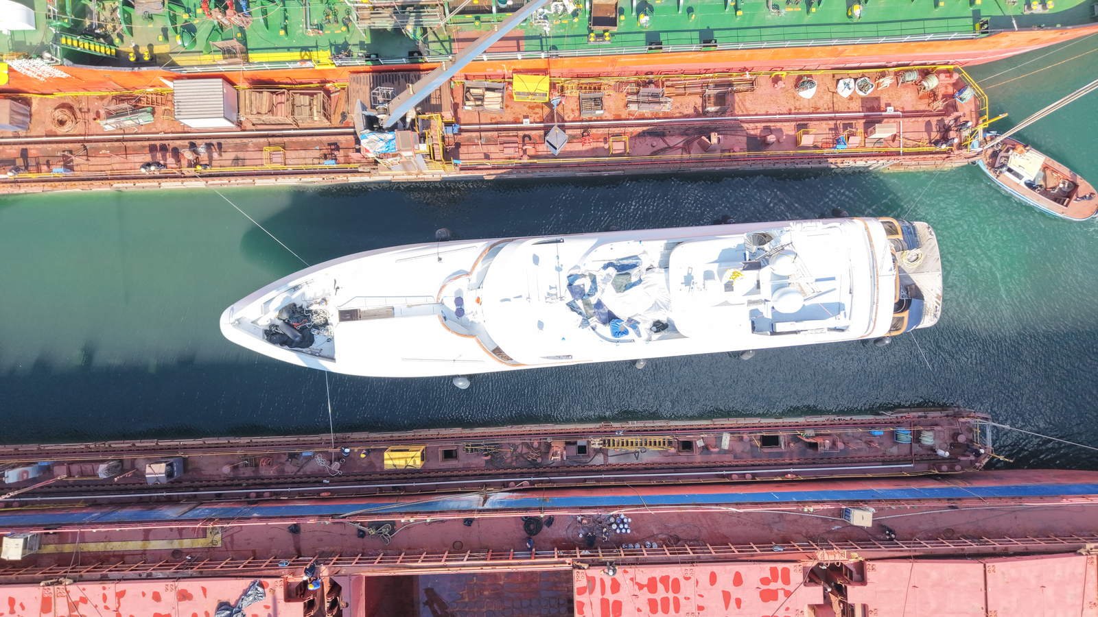 Overhead view of a white superyacht in a Tuzla repair basin with a corroded commercial hull lying along either side, plate seams and weld lines visible in the steel