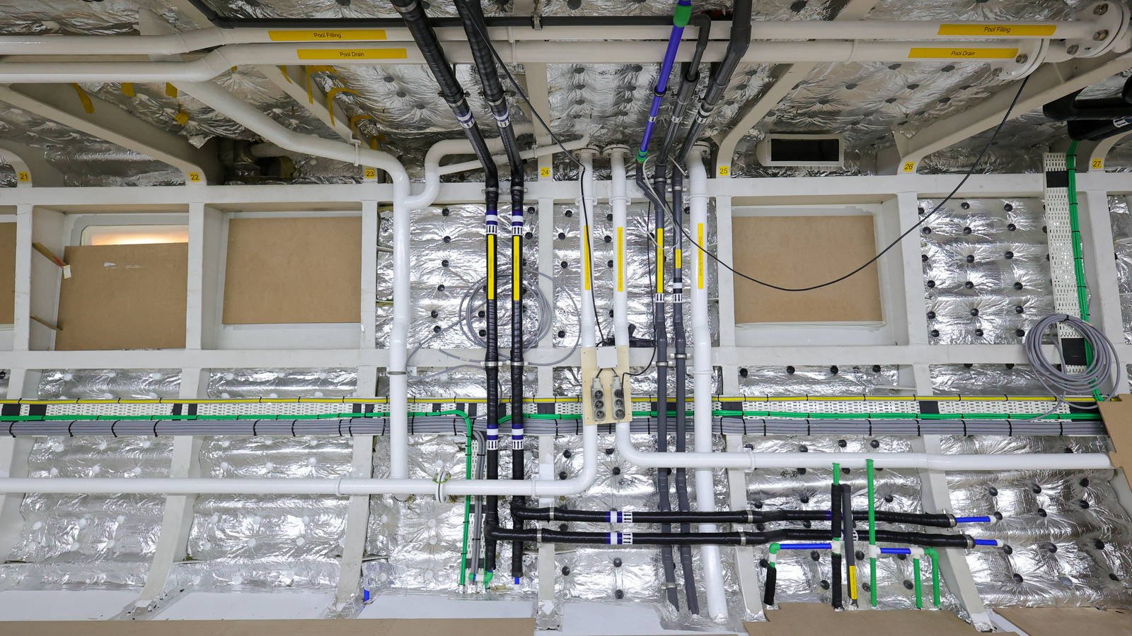 Pipe, cable and duct runs crossing an insulated deckhead in a technical space during fit-out, colour coded and labelled, with bare foil insulation and framing behind them