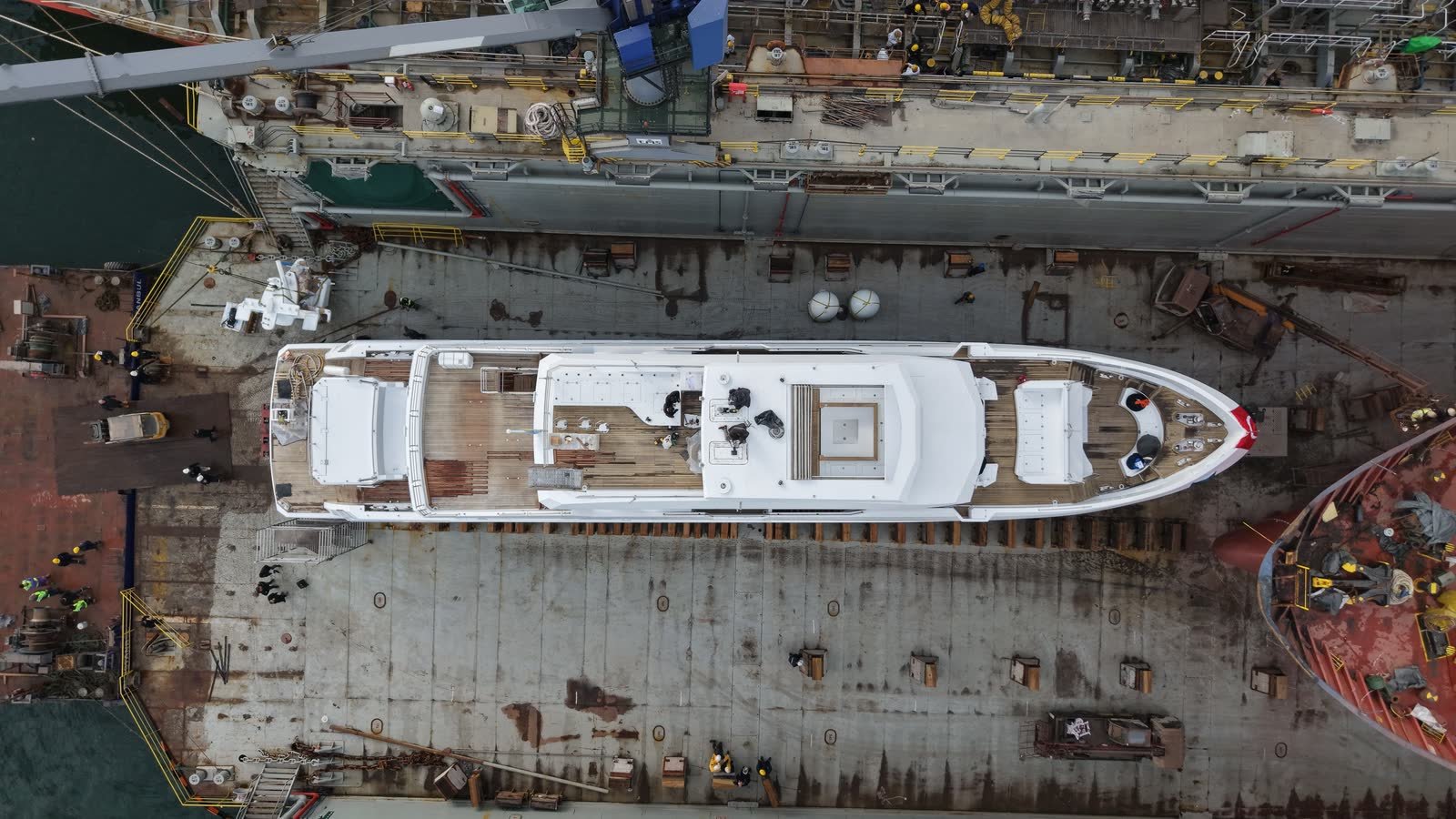A motor yacht supported on keel blocks in a dry dock during refit, seen from directly overhead, with yard hands working along both sides