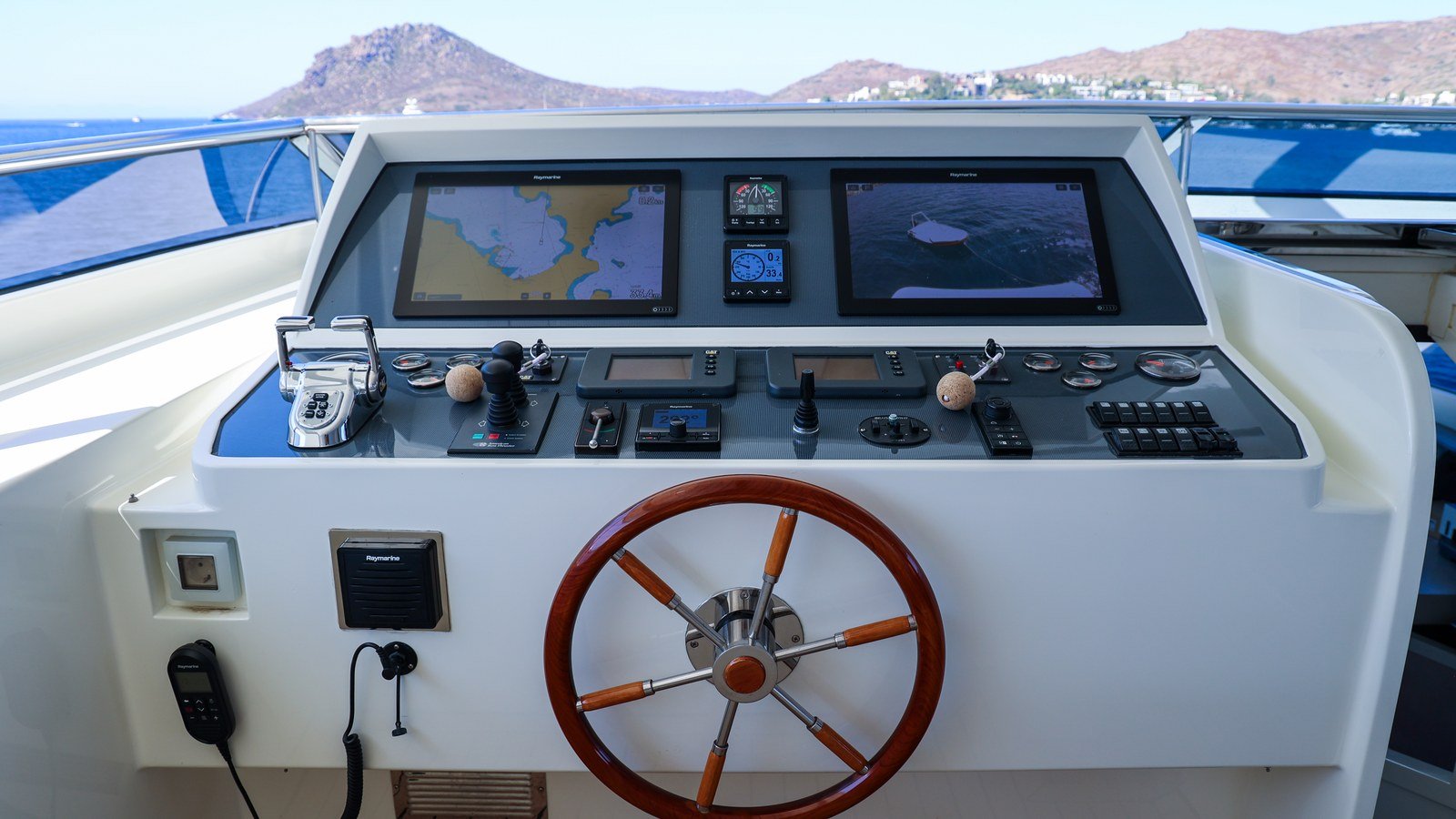 An upper helm station at sea with chart plotter and radar screens lit, twin throttles, engine instruments and a varnished wheel, open water and a coastline beyond