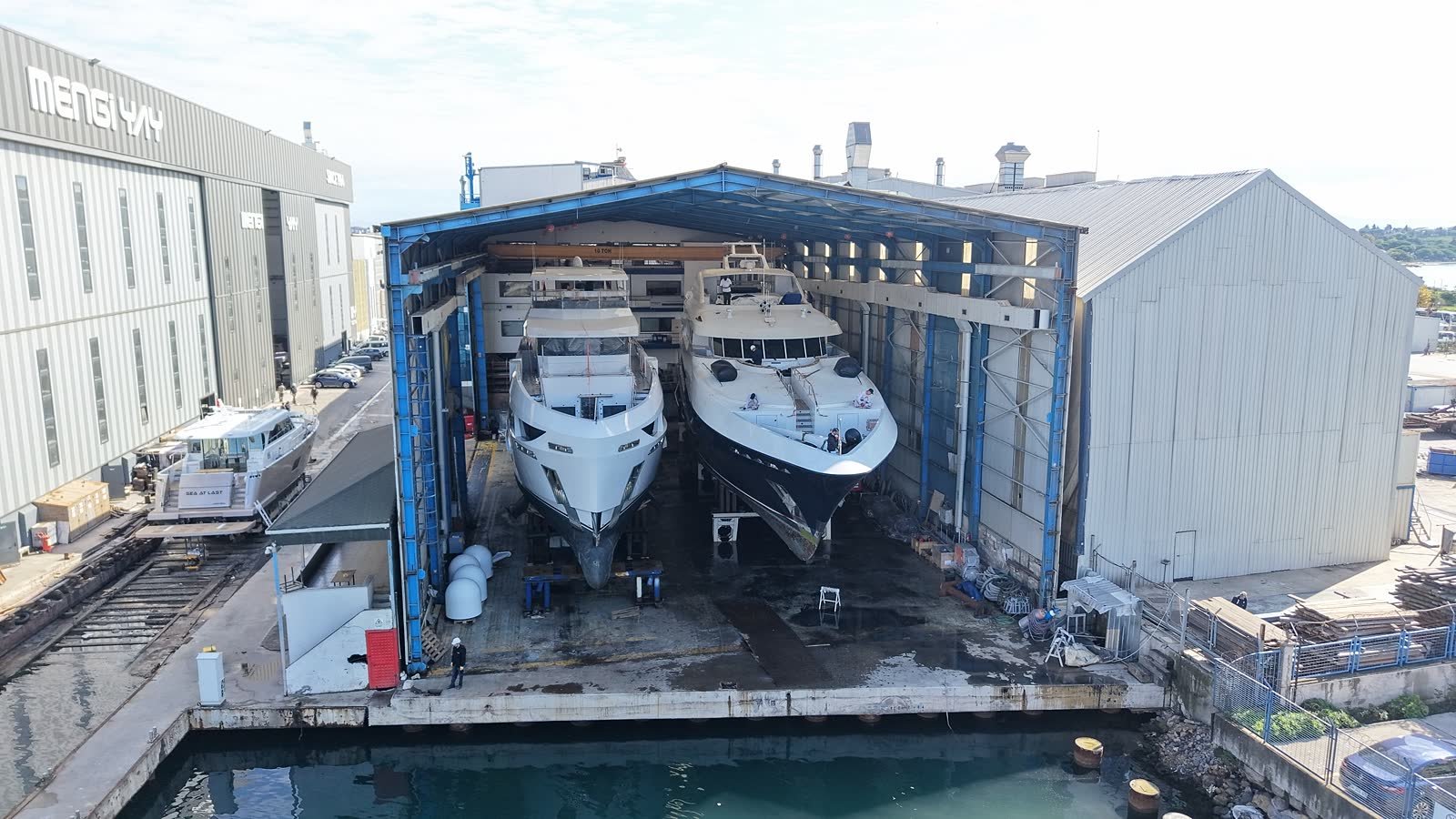 Two motor yachts standing on keel blocks and side shores inside an open-ended covered hall at the Tuzla yard, with the workshop buildings behind