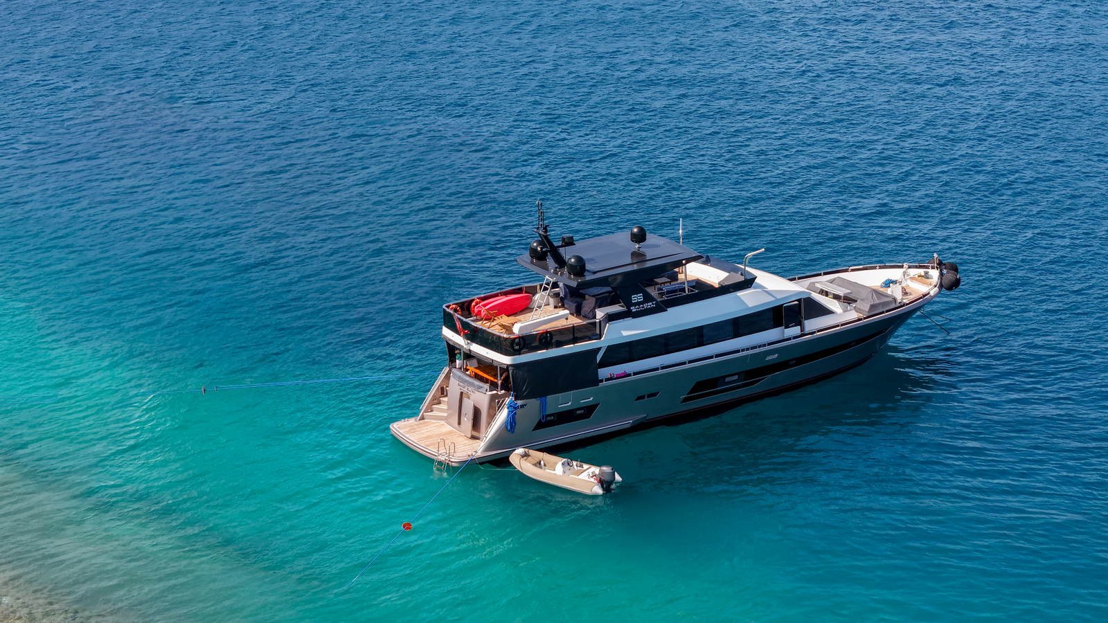 Aerial view of a motor yacht lying at anchor in clear water with her tender made fast astern and the bathing platform down