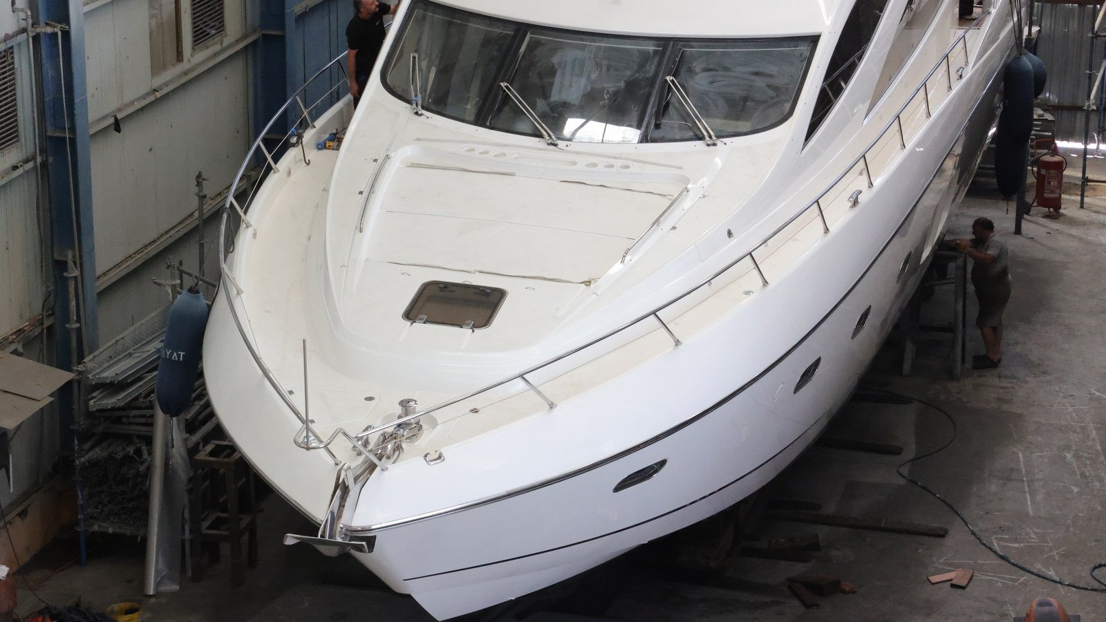 A motor yacht standing ashore on blocks and side shores inside a covered hall, her hull exposed below the waterline with fitters working alongside at hull level